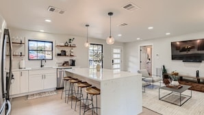 A modern kitchen with a central island, bar stools, a dining area with a sofa, and a TV mounted on the wall.