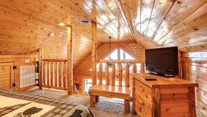 Une chambre chaleureuse aménagée dans un chalet en bois, avec un plafond en bois, un téléviseur posé sur un meuble en bois, un lit recouvert d’une couverture à motifs et une fenêtre offrant une vue agréable.