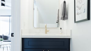Ein Badezimmer mit weißem Waschtisch, dunklen Schränken, einem goldenen Wasserhahn und einem gerahmten Kunstwerk an der Wand.