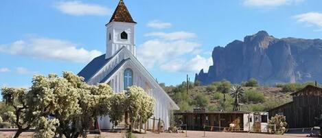 A white church with a cross on top, surrounded by desert vegetation and a mountain in the background.