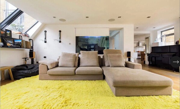 A modern living room with a sectional sofa, a large window, and a visible kitchen area.