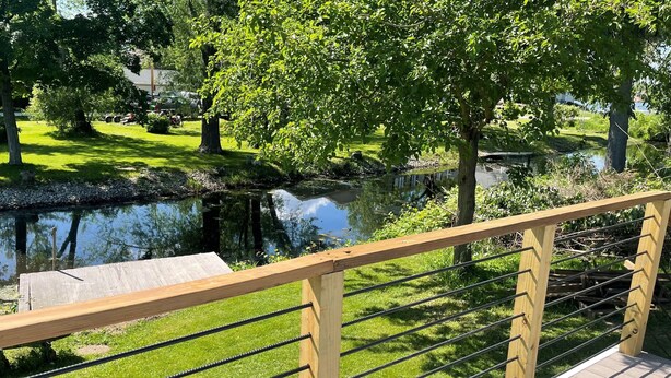 A wooden deck with a metal railing overlooking a calm pond, surrounded by lush greenery and trees. - Cottage On The Canal - An All Seasons Getaway - Waterford - Lake Tichigan (Waterford)