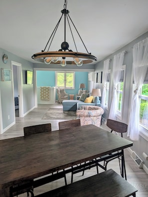 A dining area with a wooden table and benches, a living room with a blue sofa and patterned cushions, and a chandelier with exposed bulbs.