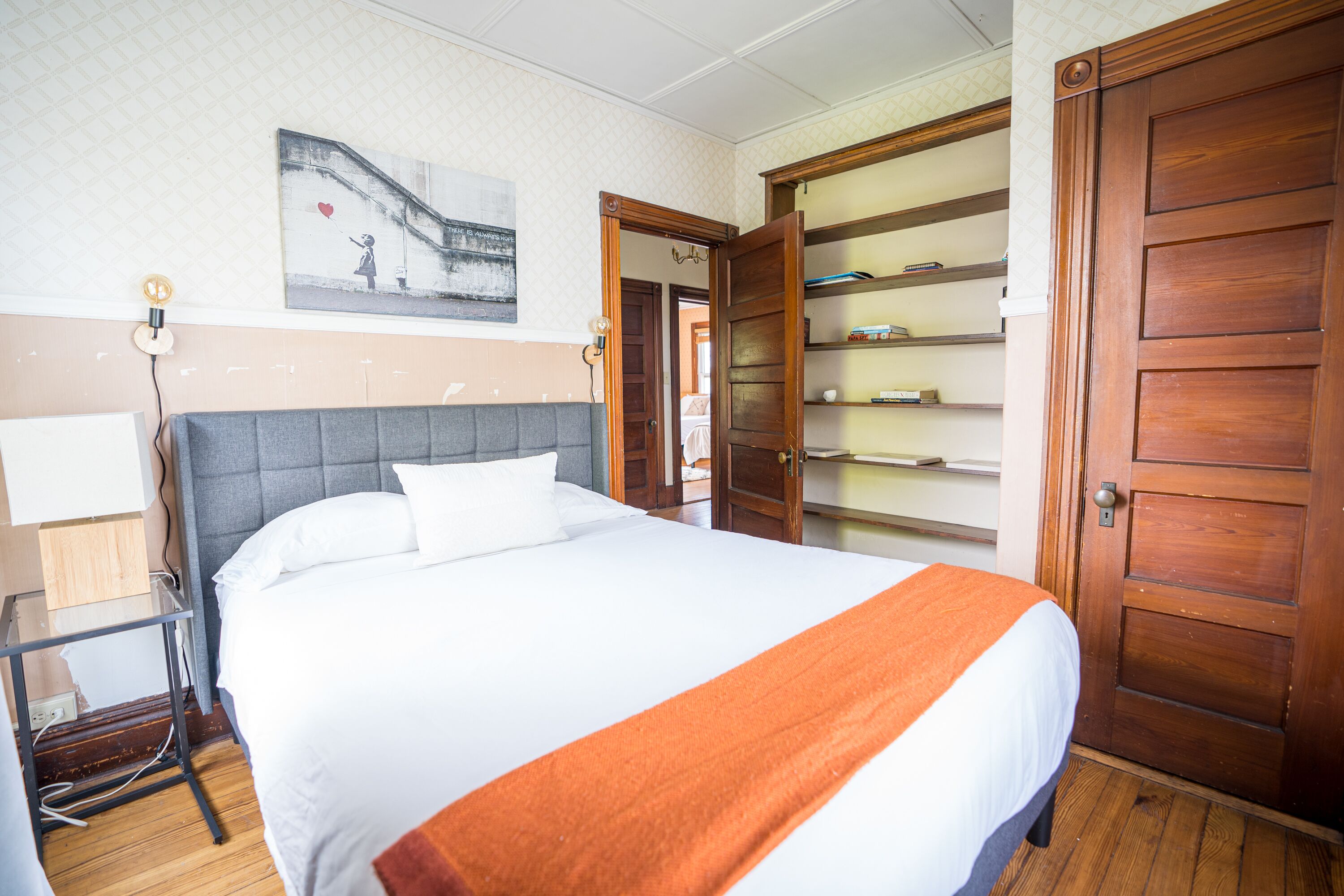 A neatly made bed with a white and orange bedspread, a grey upholstered headboard, and a wooden door to the right.