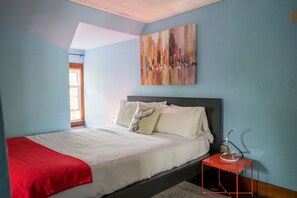 A bedroom with a bed, a red side table, a painting on the wall, and a window with a view.