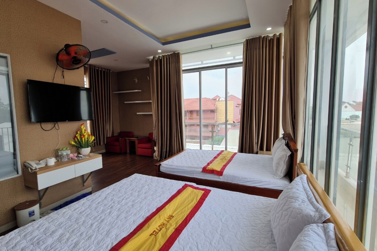 Habitación de hotel con dos camas, televisor, escritorio con teléfono y planta, y ventanas grandes con cortinas.