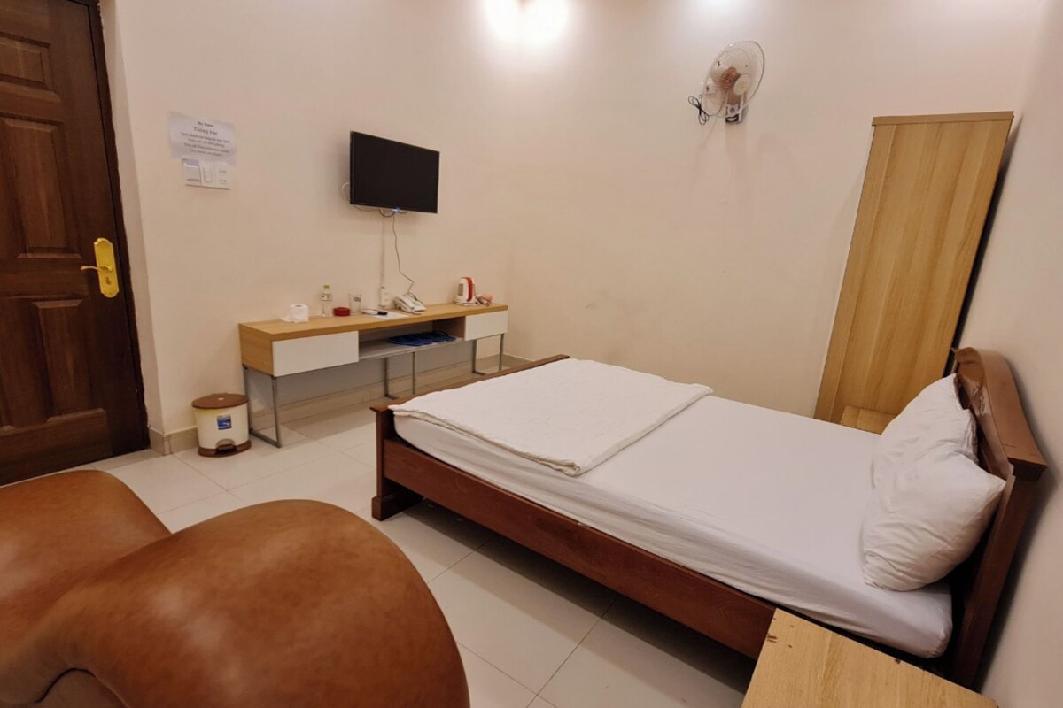 A bedroom with a wooden bed, a desk with a computer, a chair, a small trash bin, and a wall-mounted TV.