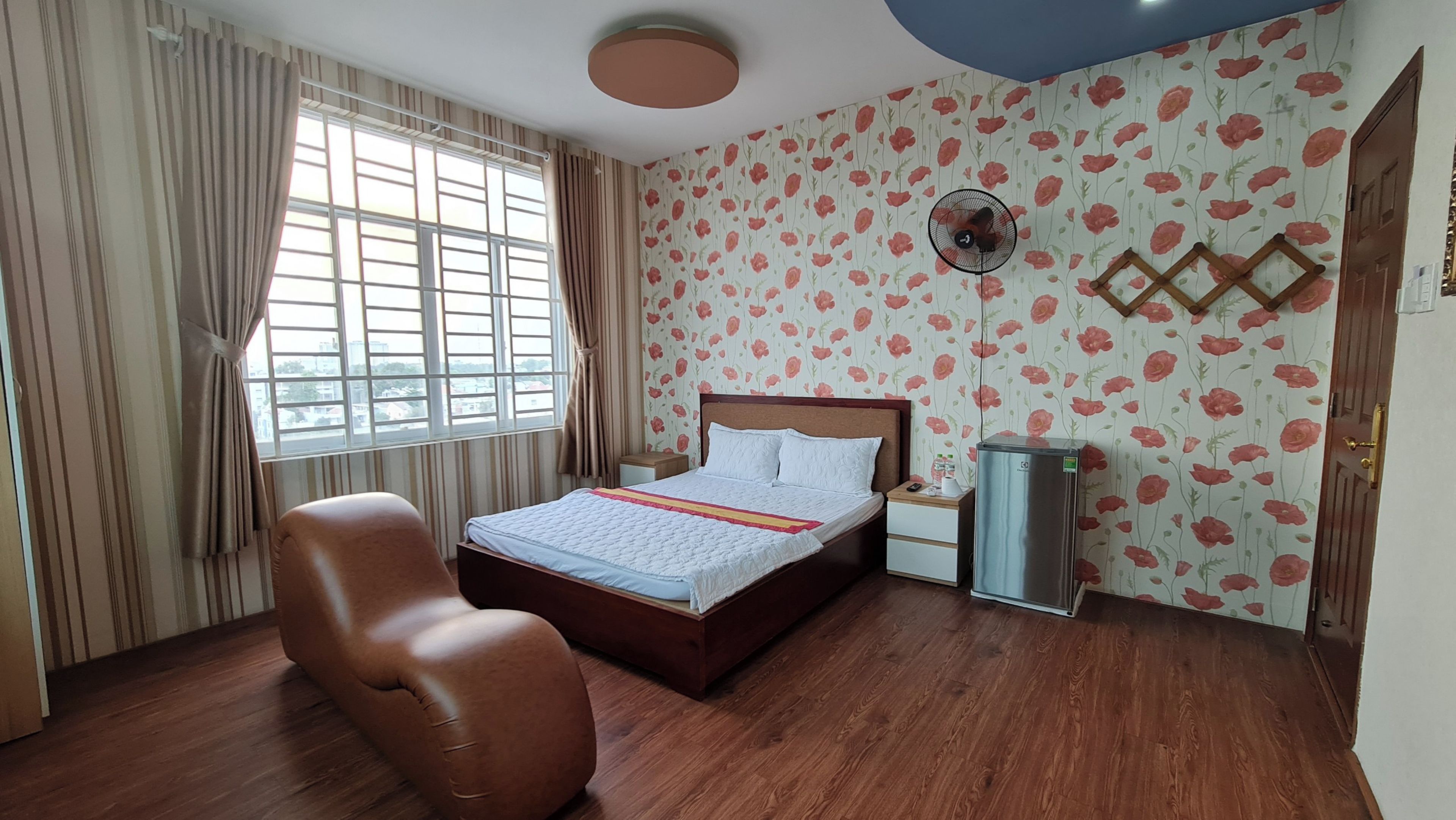 A bedroom with a bed, a chair, a bedside table, a wall-mounted fan, and a window with curtains.