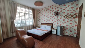 Ein Schlafzimmer mit einem Bett, einem Sessel, einem Nachttisch, einem Wandventilator und einem Fenster mit Vorhängen.