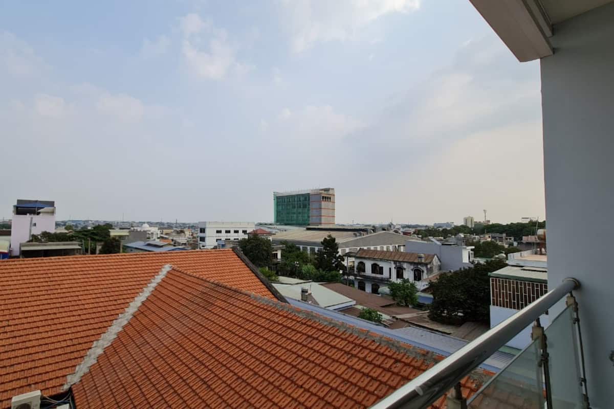 Une vue depuis un balcon, avec un toit recouvert de tuiles, donnant sur un paysage urbain composé de différents bâtiments, dont une imposante structure moderne et plusieurs bâtiments plus petits.