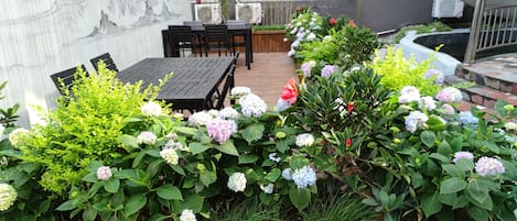 Une terrasse avec une table et des chaises en bois, entourée d’un jardin agrémenté de plantes d’hortensia.
