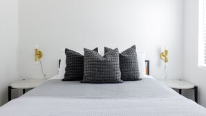Un lit bien fait, recouvert d’une couette blanche et grise, flanqué de deux tables de chevet surmontées de lampes dorées, dans une chambre minimaliste aux murs blancs.