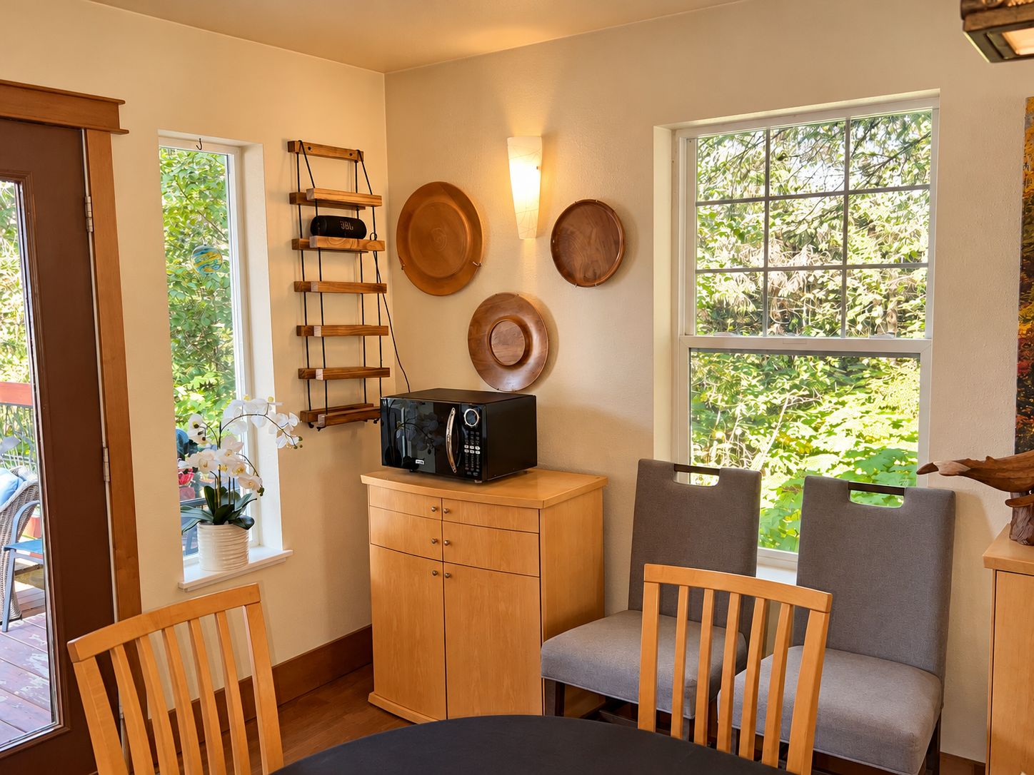 A compact kitchen with a microwave, wooden cabinets, a chair, and a window with a view of greenery.
