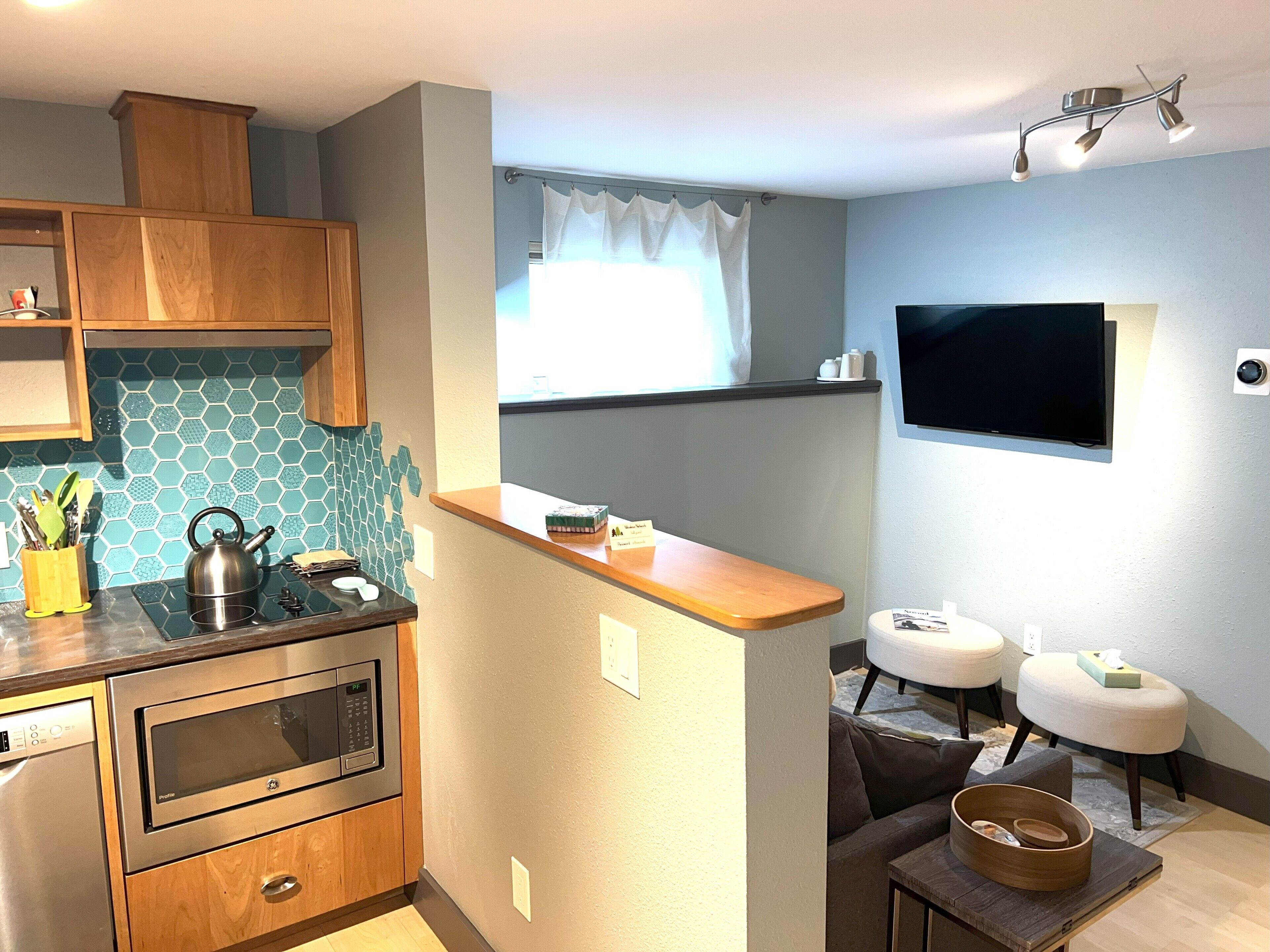 Una cocina compacta con microondas, hervidor y armarios de madera. En la zona de estar contigua hay una pequeña mesa de madera y dos taburetes.