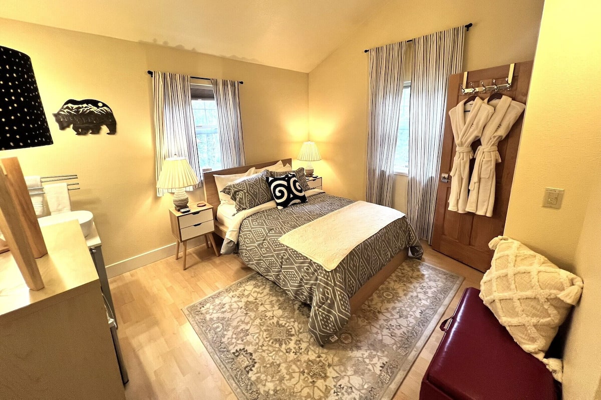 Ein Hotelzimmer mit einem Bett, zwei Nachttischen, einem Spiegel, einem Fenster mit Vorhängen, einem Teppich und einem Kleiderständer mit Bademänteln.