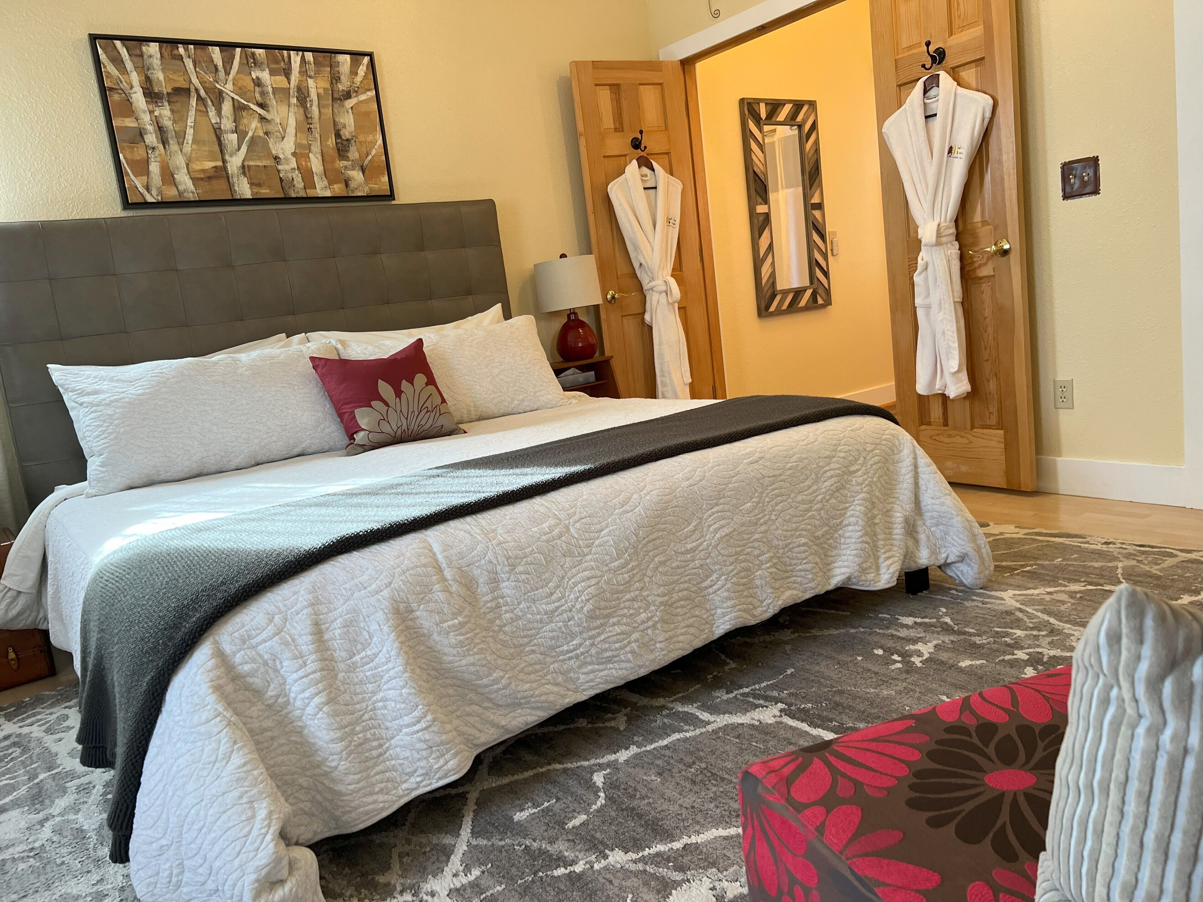 Ein ordentlich bezogenes Bett mit grauem Kopfteil, weißen und roten Kissen und einer grauen und weißen Decke. Zwei Holztüren mit weißen Bademänteln, die an Haken hängen, und ein gerahmtes abstraktes Gemälde über dem Bett.