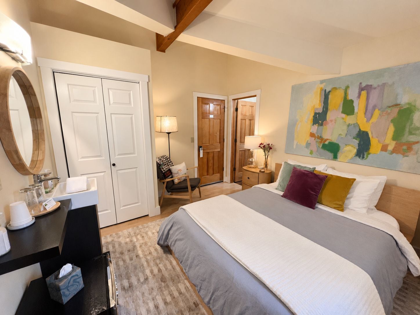 A hotel room with a bed, bedside table, lamp, chair, and a painting on the wall.