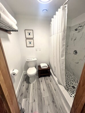 A bathroom with a toilet, a small wooden shelf with towels, a shower with a marble wall, and a wooden floor. - Bell In The Woods B&B (Seward)