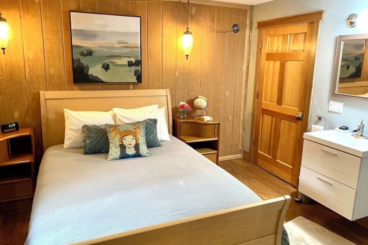 Ein Schlafzimmer mit einem hölzernen Kopfteil, einem Bett mit weißer Bettwäsche, einem Nachttisch, einem Beistelltisch, einem Holzschrank und einem Spiegel an der Wand.
