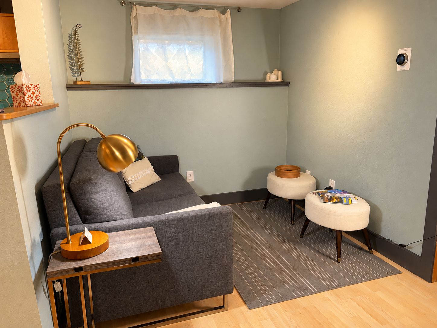 Un salon moderne comprenant un canapé, une table d’appoint, une lampe, un petit tabouret et une fenêtre avec un rideau.