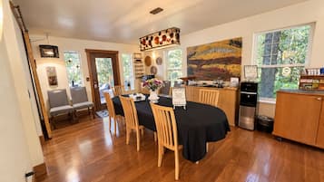 A dining area with a black tablecloth, wooden chairs, a vase with flowers, and a kitchen area with a refrigerator and a coffee machine.