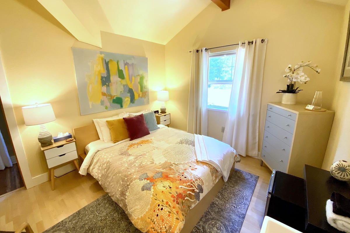 Ein Schlafzimmer mit einem großen Bett, einem Nachttisch, einer Kommode, einem Fenster mit Vorhängen und einem Gemälde an der Wand.