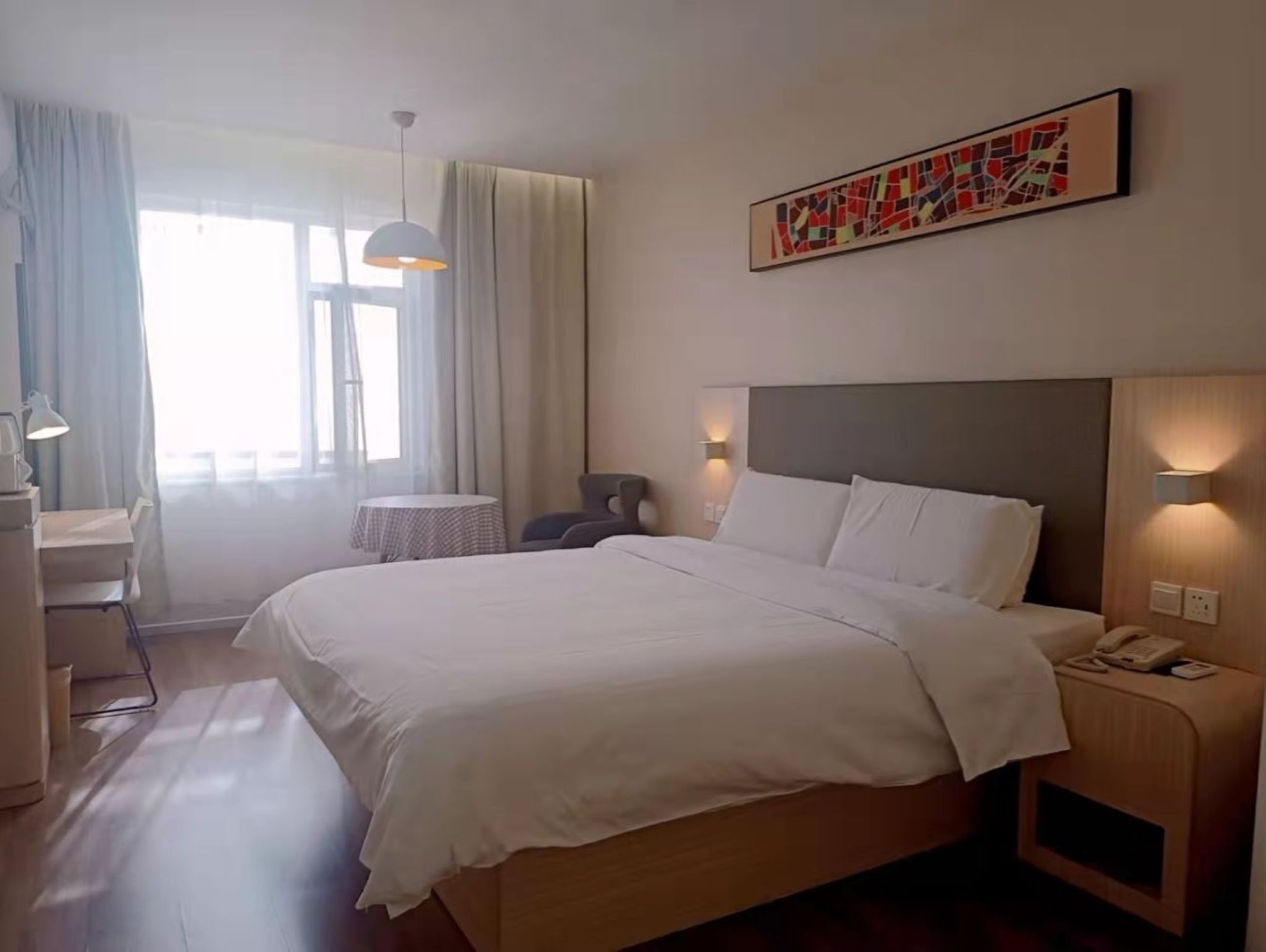 A hotel room with a bed, bedside tables, a desk, and a chair. There is a window with curtains, a hanging light fixture, and a painting on the wall.