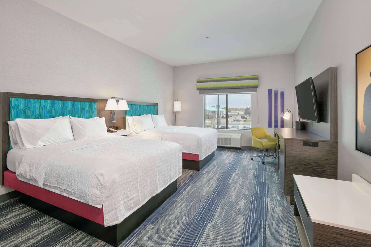 A hotel room with two beds, a desk, a chair, a TV, and a window with a view of the city.