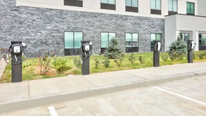 Een modern gebouw met een gevel van natuursteen, veel ramen en oplaadpunten voor elektrische voertuigen langs de stoep.