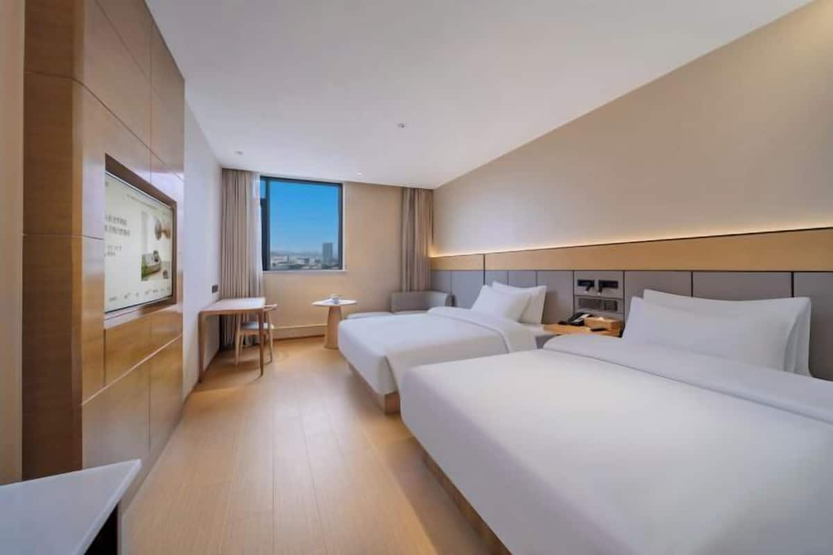 A hotel room with two beds, a desk, a chair, a small table, and a large window with a city view.