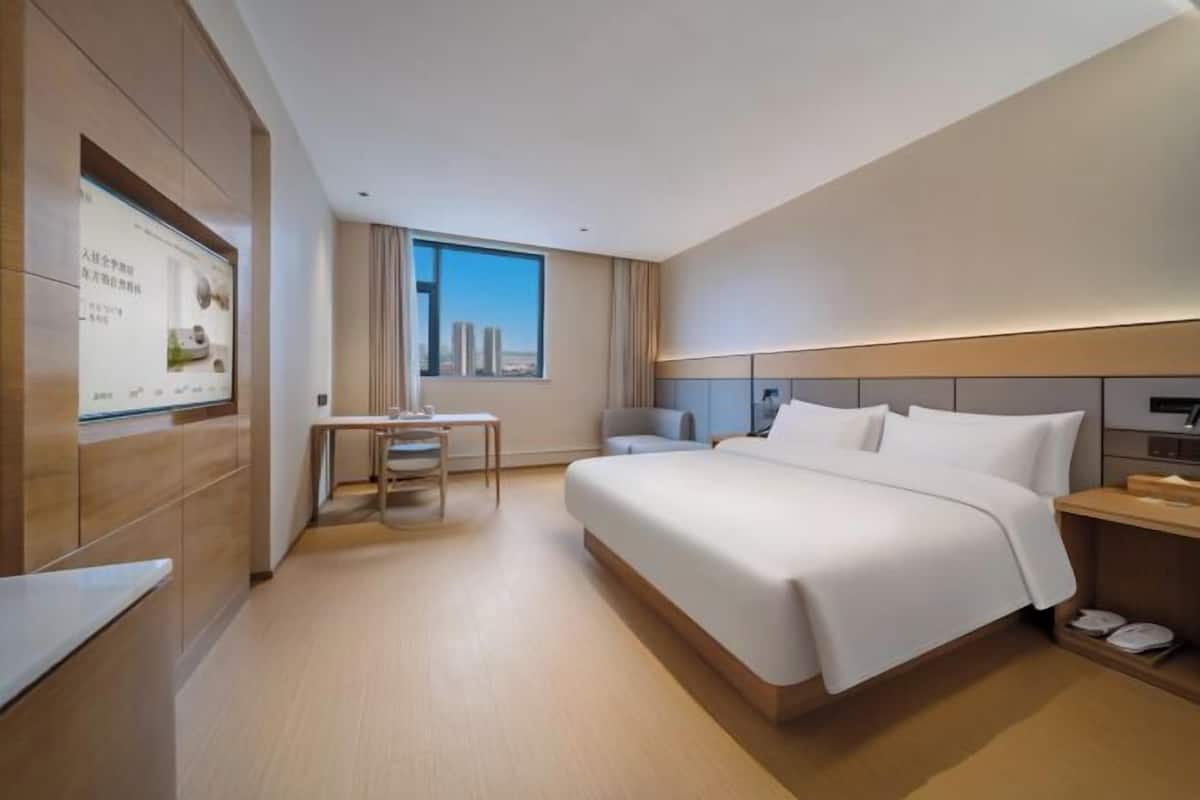A modern hotel room with a large bed, a desk, a chair, and a view of the city.