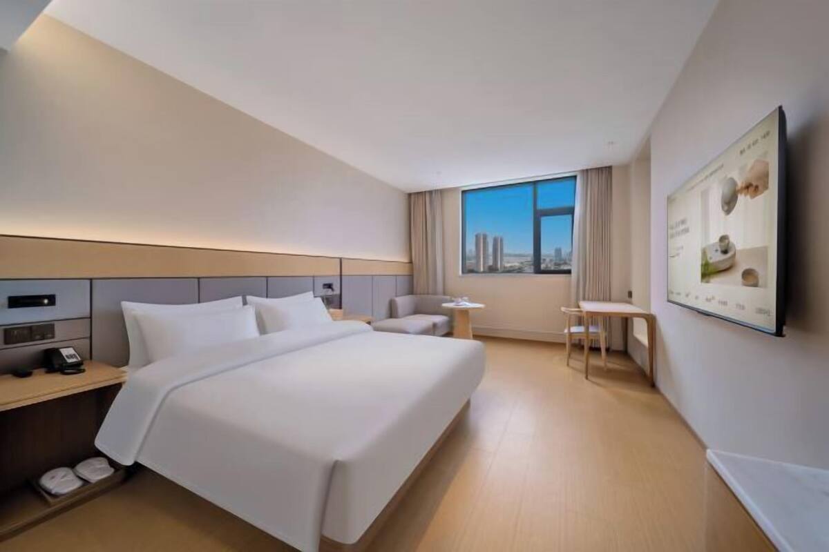 A modern hotel room with a bed, bedside table, chair, and a large window with a city view.