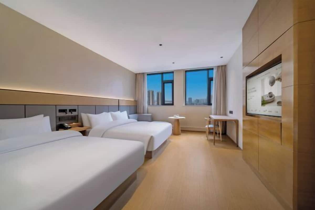 A hotel room with two beds, a small table, a chair, a TV mounted on the wall, and a city view through the windows.