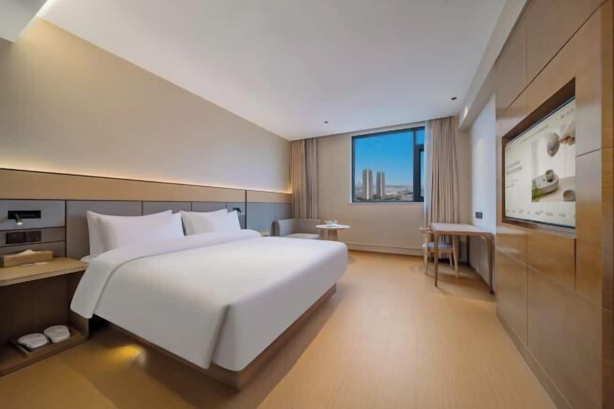 A modern hotel room with a large bed, a desk, and a view of buildings.