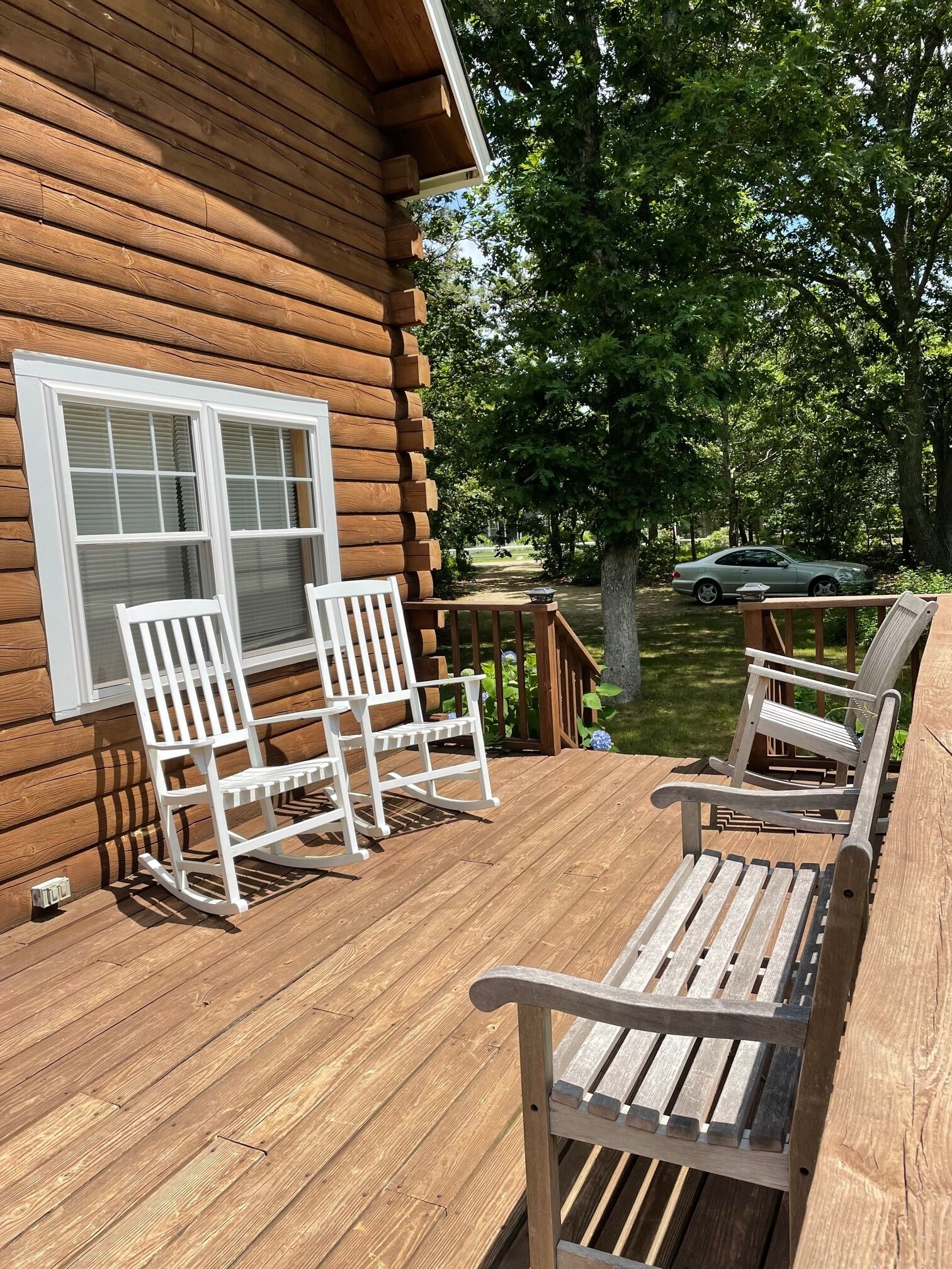 Eine Holzterrasse mit zwei Schaukelstühlen und einem Tisch, mit Blick auf einen Garten, in dem sich ein Auto befindet.