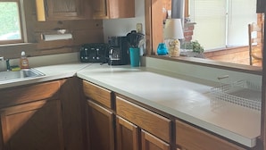 Une cuisine avec des meubles de rangement en bois, un plan de travail blanc, un évier et une fenêtre avec des stores.