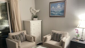 Un salon avec deux fauteuils, une table basse en verre, une commode blanche, un tableau et une lampe.