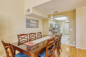 A dining area with a wooden table and chairs, a painting on the wall, and a kitchen in the background.