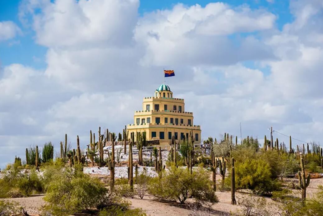 A large, multi-story building with a distinctive dome and flag, surrounded by desert vegetation.
