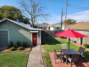 Un jardin avec une terrasse en briques, un parasol rouge et une table entourée de chaises.
