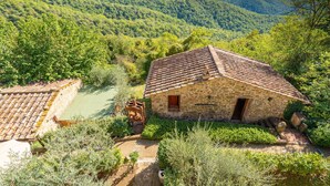 Un bâtiment en pierre, surmonté d’un toit de tuiles, entouré d’une végétation luxuriante et de collines.