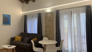 Ein modernes Wohnzimmer mit einer Couch, einem kleinen runden Tisch und zwei Stühlen. Eine Wand aus Stein bildet einen Blickfang und ein großes Fenster ist mit Vorhängen versehen.