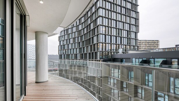 Un bâtiment moderne doté d’une façade en verre incurvé, d’une terrasse en bois et d’une rambarde en métal.