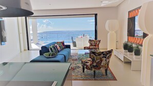 Un salón moderno con un ventanal que ofrece vistas al mar, un sofá azul con cojines estampados, un sillón con diseño y un televisor de pantalla plana montado en la pared.