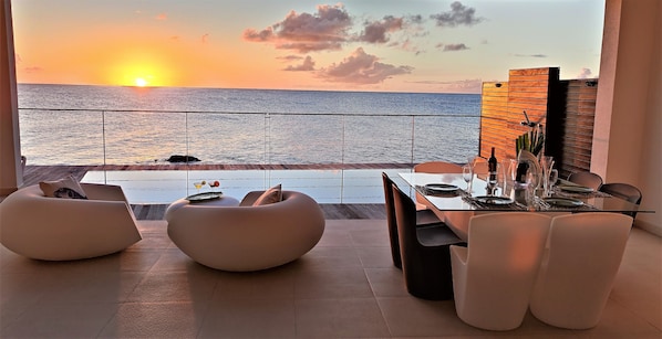 Un espace de restauration extérieur moderne, offrant une vue sur le coucher de soleil sur l’océan, avec un mobilier contemporain et une table en verre.