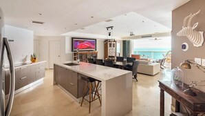 Een moderne keuken met een centraal eiland, een eethoek met uitzicht op de oceaan, en een woonruimte met een grote televisie.