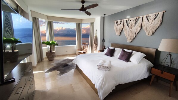 A spacious bedroom with a large bed, a ceiling fan, a view of the ocean, and a macrame wall hanging.