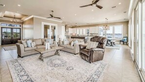 A spacious living area with a large sofa set, a coffee table, and a fireplace. There are ceiling fans and a view of the outside through large windows.