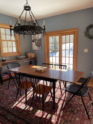 Une salle à manger avec une table en bois, des chaises noires, un lustre et une grande fenêtre donnant sur un paysage enneigé.