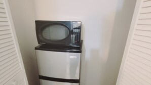 Un petit réfrigérateur, surmonté d’un four à micro-ondes, est installé dans un couloir étroit aux murs blancs et au sol carrelé.